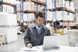A young man using his laptop and checking his notes while working in a warehouse